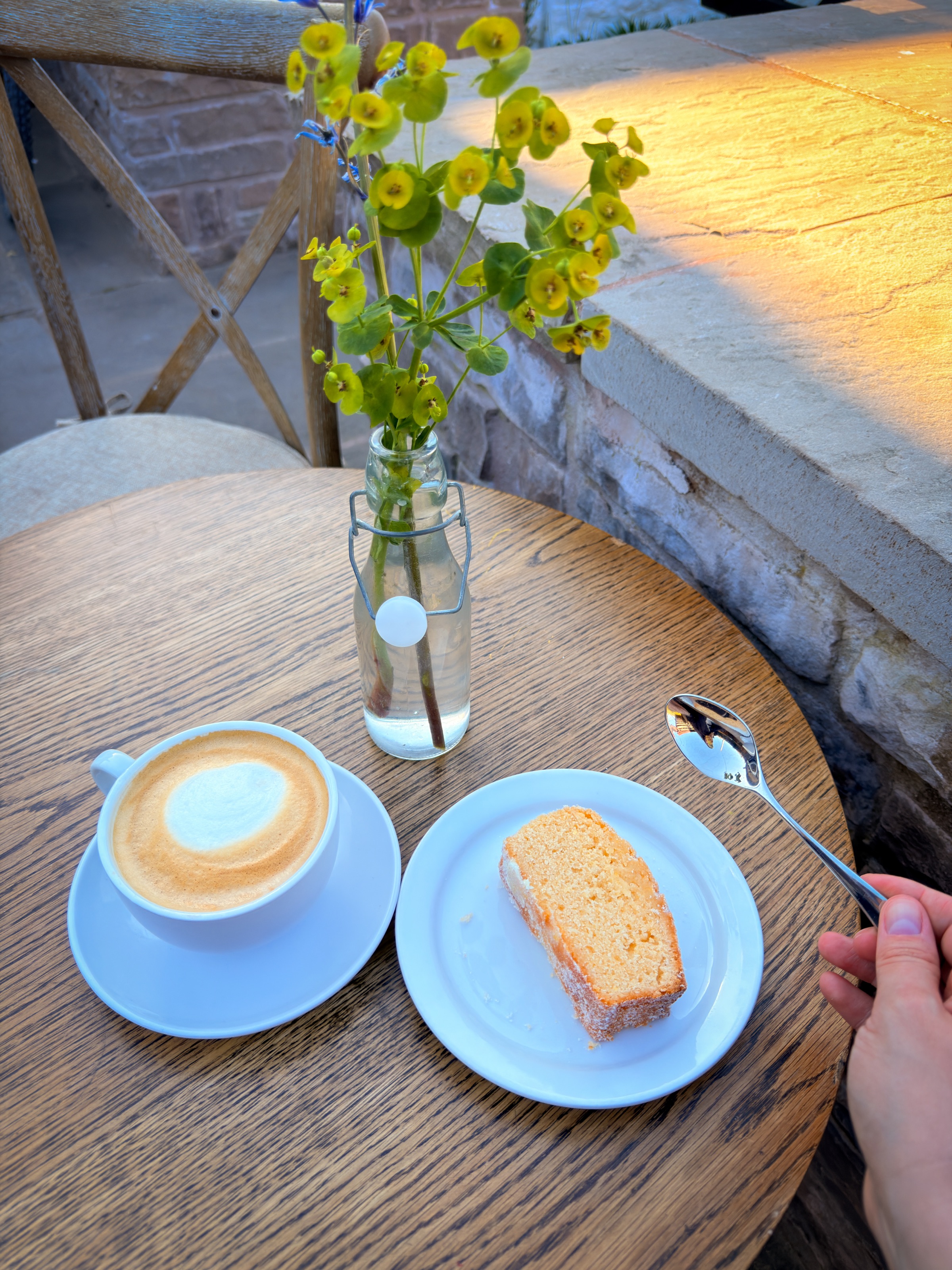 A flat white and a slice of lemon drizzle on an outdoor table at Godric's, with a glass jar of yellow wildflowers and the warm sandstone of the building behind.