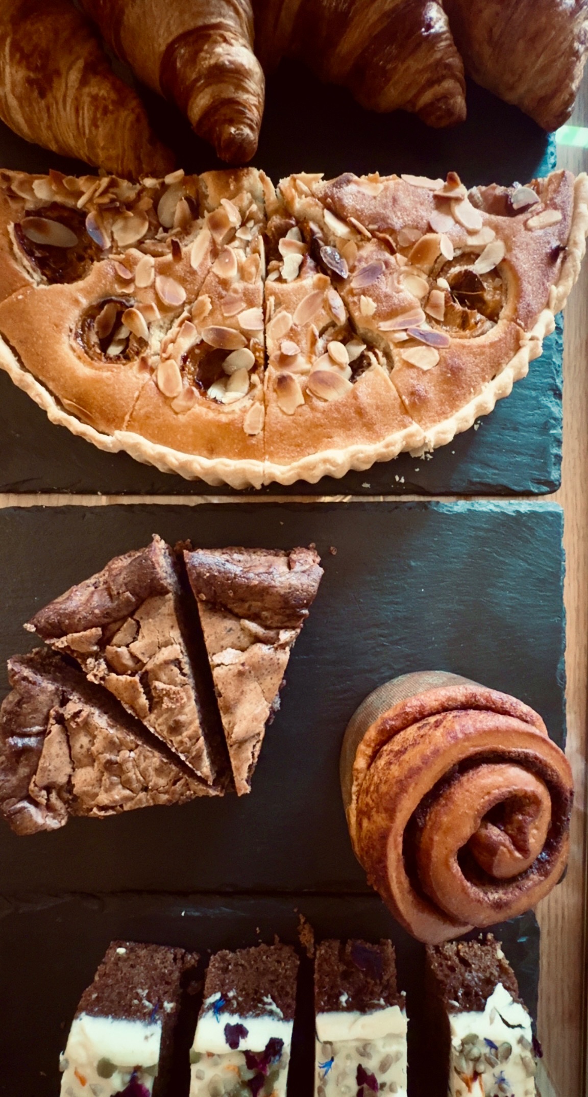 A slate counter laid with croissants, an almond frangipane tart, brownies, a cinnamon bun and slices of layered cake.